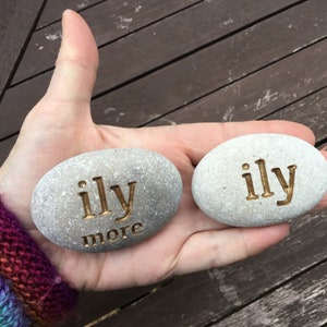 May include: Two gray, oval-shaped stones with the words "ily" and "more" engraved in a gold color. The stones are held in a hand, with a wooden surface in the background. The text is a declaration of love.