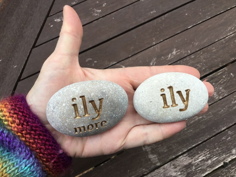 May include: Two gray, oval-shaped stones with the words "ily" and "more" engraved in a gold color. The stones are held in a hand, with a wooden surface in the background. The text is a declaration of love.