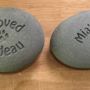 May include: Two gray, smooth, oval-shaped stones with engraved messages. The first stone says "Beloved Cadeau" with a paw print. The second stone says "Mia's Tree" with a paw print.