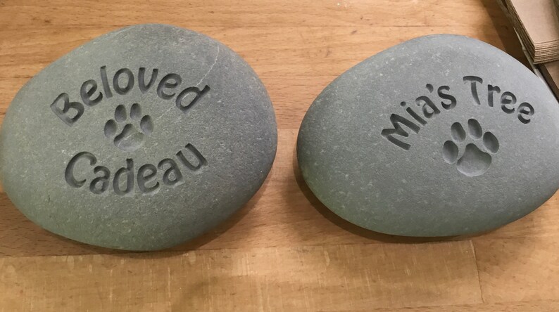 May include: Two gray, smooth, oval-shaped stones with engraved messages. The first stone says "Beloved Cadeau" with a paw print. The second stone says "Mia's Tree" with a paw print.