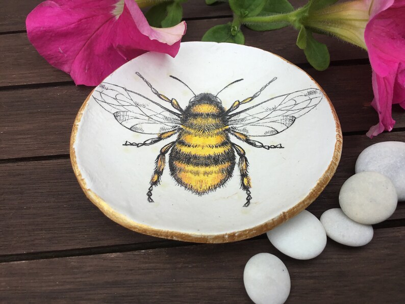 Op de afbeelding: Een witte keramische schaal met een gouden rand heeft een gedetailleerde illustratie van een hommel met zwarte en gele strepen. De schaal staat op een houten oppervlak.