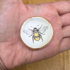 May include: A small white ceramic dish with a gold rim features a hand-painted image of a bee with black and yellow stripes. The bee is centered on the dish and has its wings spread out.