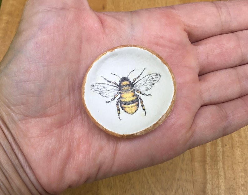 May include: A small white ceramic dish with a gold rim features a hand-painted image of a bee with black and yellow stripes. The bee is centered on the dish and has its wings spread out.