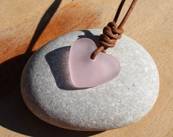 Pode incluir: Um pingente de coração de vidro rosa em um cordão de couro marrom. O pingente está apoiado em uma pedra cinza.