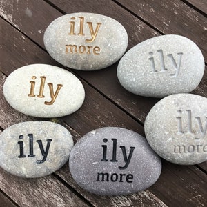 May include: Six gray river stones with the engraved text "ily" or "ily more" in various colors. The stones are arranged on a dark brown wooden surface. The stones are a decorative item.