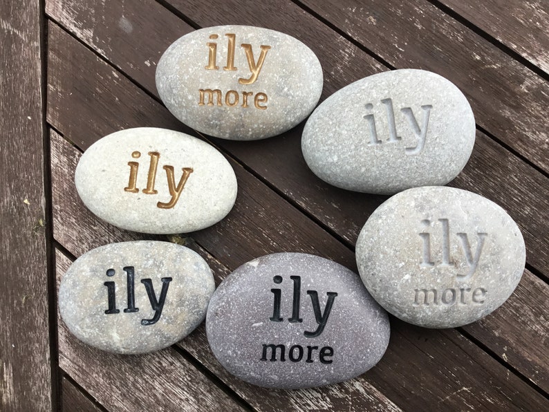 May include: Six gray river stones with the engraved text "ily" or "ily more" in various colors. The stones are arranged on a dark brown wooden surface. The stones are a decorative item.