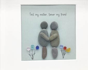 Puede incluir: Una pieza de arte de guijarros enmarcada que representa a dos figuras de pie una al lado de la otra con los brazos alrededor. Las figuras están hechas de guijarros grises y marrones. El texto "First my mother, forever my friend" está escrito sobre las figuras. Hay pequeños guijarros de vidrio de colores dispuestos en forma de flor a cada lado de las figuras.