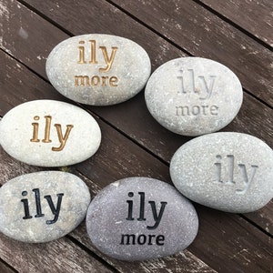 May include: Six smooth, gray river stones with the words "ily" and "ily more" engraved in various colors. The stones are arranged on a dark wooden surface, creating a natural and heartfelt display.