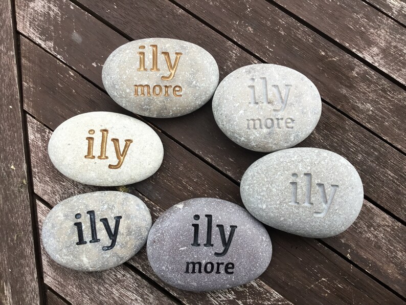 May include: Six smooth, gray river stones with the words "ily" and "ily more" engraved in various colors. The stones are arranged on a dark wooden surface, creating a natural and heartfelt display.
