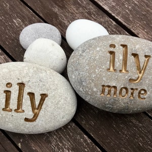 May include: Two gray river stones with the words "ily" and "ily more" carved into them. The stones are surrounded by other smooth, gray and white stones. The background is a dark wooden surface.
