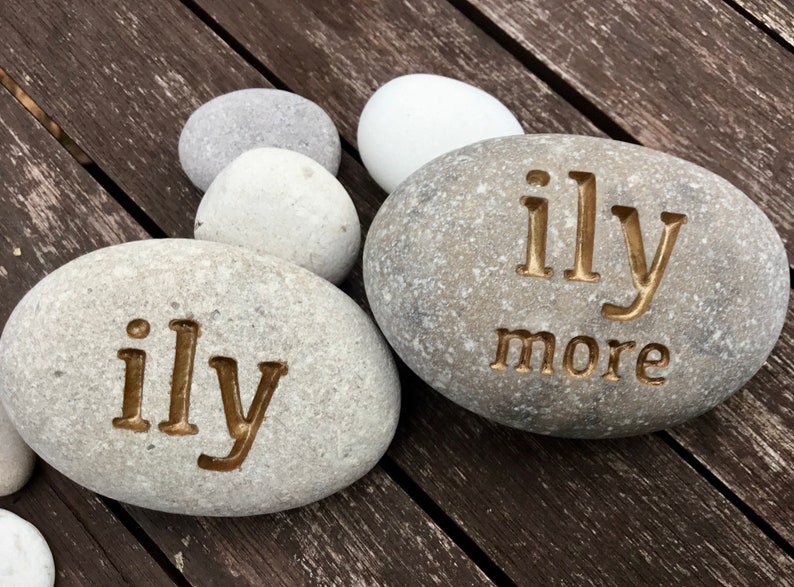 May include: Two gray river stones with the words "ily" and "ily more" carved into them. The stones are surrounded by other smooth, gray and white stones. The background is a dark wooden surface.