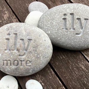 May include: Two gray river stones with the carved text "ily" on each. One stone also has "more" carved below. Additional smaller stones are visible. The stones are on a weathered wooden surface.