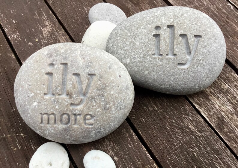 May include: Two gray river stones with the carved text "ily" on each. One stone also has "more" carved below. Additional smaller stones are visible. The stones are on a weathered wooden surface.