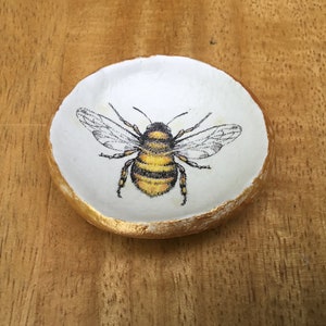 May include: A small white ceramic dish with a gold rim, featuring a hand-painted image of a bumblebee.