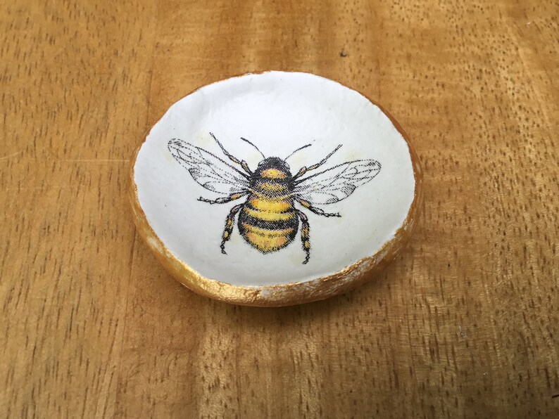 May include: A small white ceramic dish with a gold rim, featuring a hand-painted image of a bumblebee.