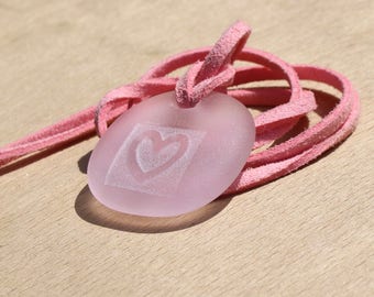 Pode incluir: Um pingente rosa oval com um coração gravado dentro de um quadrado. O pingente está preso a um cordão de camurça rosa. O colar é exibido em uma superfície de madeira marrom clara.