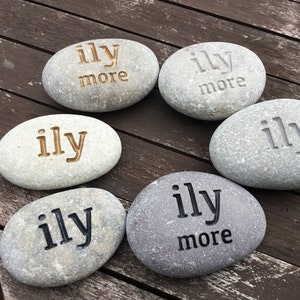 May include: Six gray river stones with the words "ily" and "ily more" engraved in various colors. The stones are arranged in a circular pattern on a wooden surface. The text is in gold, black, and gray.