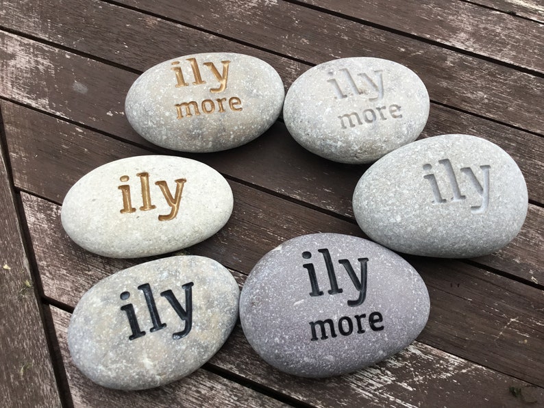May include: Six gray river stones with the words "ily" and "ily more" engraved in various colors. The stones are arranged in a circular pattern on a wooden surface. The text is in gold, black, and gray.