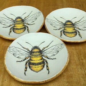 Op de afbeelding: Drie witte keramische schalen met gouden rand, elk met een afbeelding van een zwarte en gele hommel.