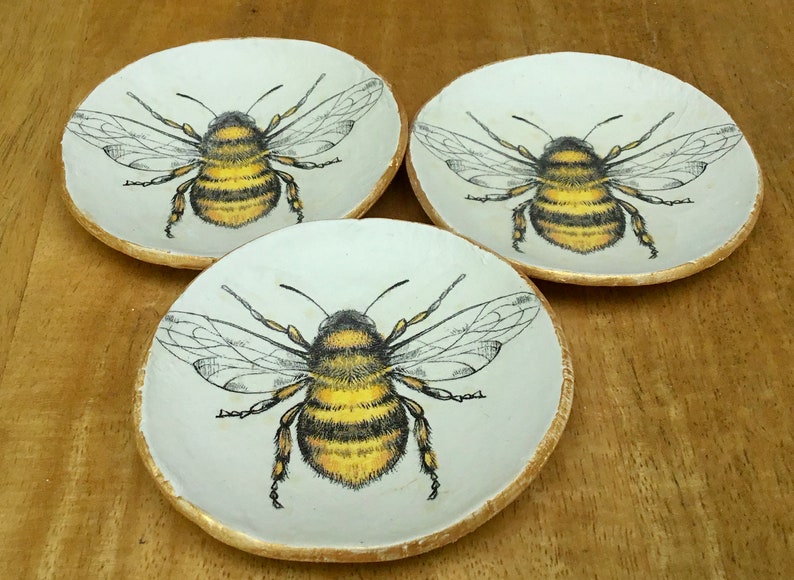 Op de afbeelding: Drie witte keramische schalen met gouden rand, elk met een afbeelding van een zwarte en gele hommel.