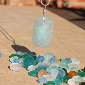 May include: A silver chain necklace with a light blue glass pendant. The pendant is shaped like a rectangle and has a frosted finish. The necklace is laying on a surface with a scattering of small, blue, green, and white glass stones.