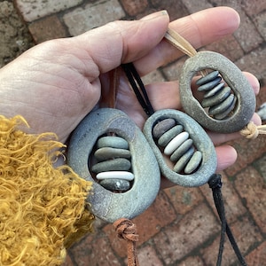 Puede incluir: Tres colgantes de piedra gris, cada uno con una pila de pequeñas piedras dentro de una abertura ovalada. Los colgantes cuelgan de cuero o cordón negro. El fondo es un entorno exterior borroso.