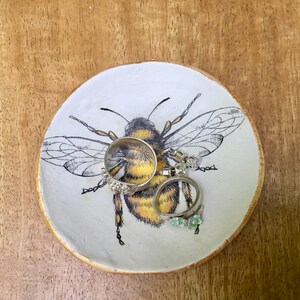 Op de afbeelding: Een witte keramische ringenschotel met een zwarte en gele bij-illustratie. De schotel heeft twee zilveren ringen en een paar oorbellen erop.