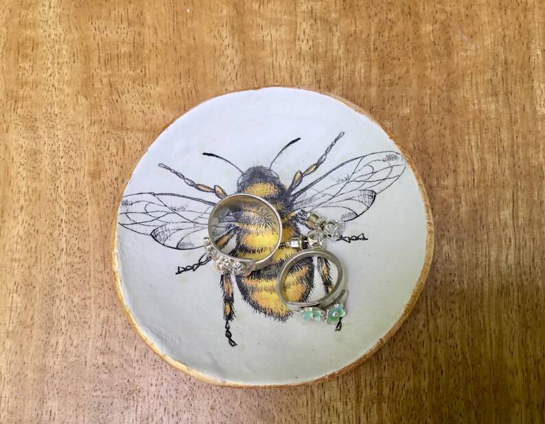 Op de afbeelding: Een witte keramische ringenschotel met een zwarte en gele bij-illustratie. De schotel heeft twee zilveren ringen en een paar oorbellen erop.