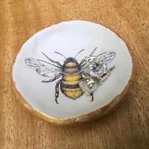 May include: A white ceramic ring dish with a gold rim, featuring a black and yellow bee illustration. Two clear crystal earrings are resting on the dish.
