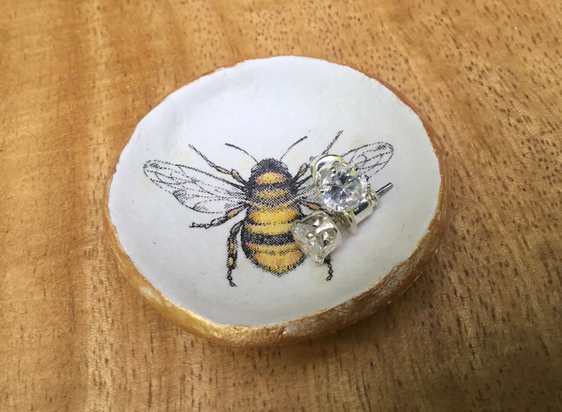 May include: A white ceramic ring dish with a gold rim, featuring a black and yellow bee illustration. Two clear crystal earrings are resting on the dish.