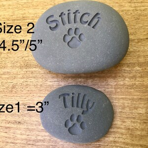 May include: Two gray engraved pet memorial stones. The larger stone is 4.5 inches by 5 inches and says "Stitch" with a paw print. The smaller stone is 3 inches and says "Tilly" with a paw print.