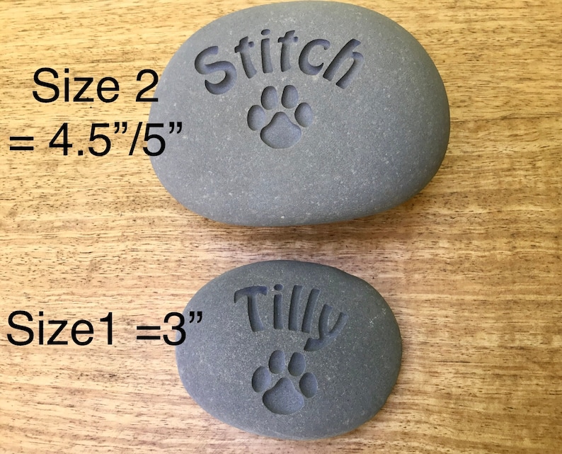 May include: Two gray engraved pet memorial stones. The larger stone is 4.5 inches by 5 inches and says "Stitch" with a paw print. The smaller stone is 3 inches and says "Tilly" with a paw print.