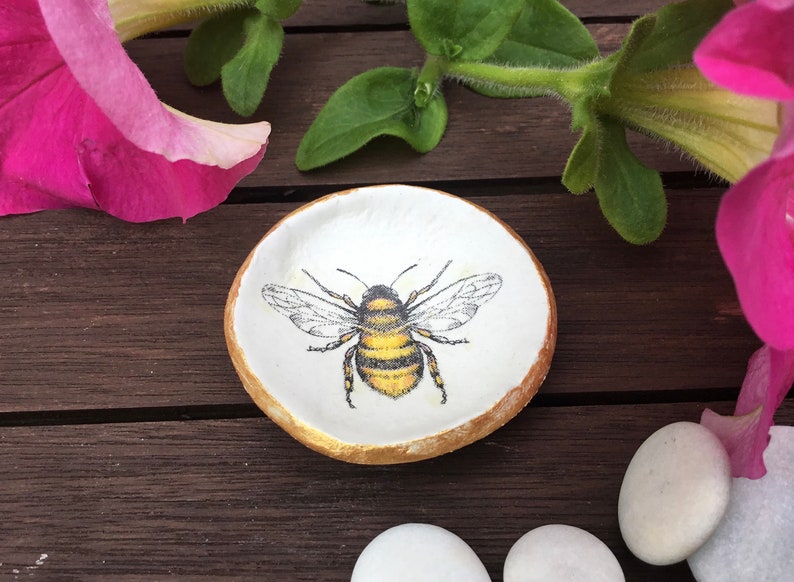 May include: A small white ceramic dish with a gold rim, featuring a hand-painted image of a bumblebee. The dish is perfect for holding small items like rings or earrings.
