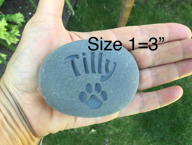May include: A gray, smooth, oval-shaped stone with the name "Tilly" and a paw print engraved on it. The stone is being held in a hand, and the text "Size 1 = 3" is visible above the stone.