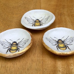 May include: Three small white ceramic dishes with gold rims. Each dish features a black and yellow bee illustration.