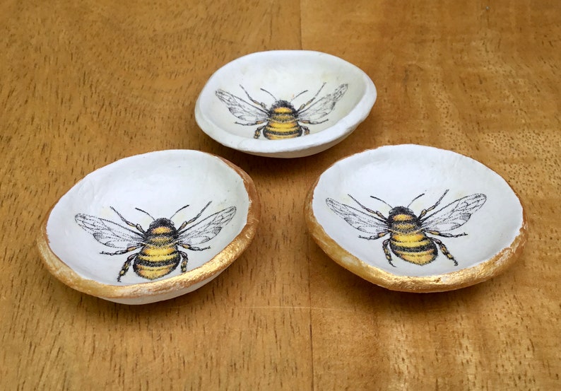May include: Three small white ceramic dishes with gold rims. Each dish features a black and yellow bee illustration.