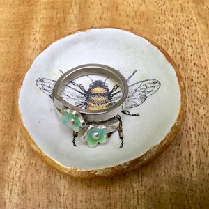 May include: A white ceramic ring dish with a gold rim and a bee design. The dish has two small green flowers on it and a silver ring is resting on the dish.