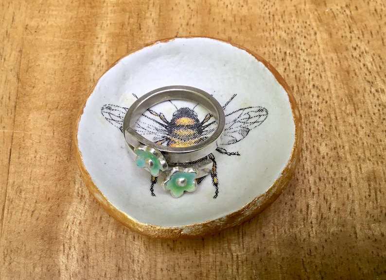 May include: A white ceramic ring dish with a gold rim and a bee design. The dish has two small green flowers on it and a silver ring is resting on the dish.