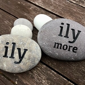 May include: Two gray river rocks with the words "ily" and "ily more" etched in black. The rocks are arranged on a weathered wooden surface, with several other smooth stones in the background. The rocks are a natural, rustic decorative item.