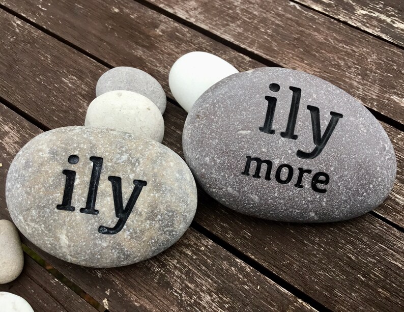 May include: Two gray river rocks with the words "ily" and "ily more" etched in black. The rocks are arranged on a weathered wooden surface, with several other smooth stones in the background. The rocks are a natural, rustic decorative item.