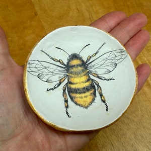 Op de afbeelding: Een wit keramiek sieradenschotel met een gouden rand heeft een handgeschilderde illustratie van een hommel met zwarte en gele strepen.