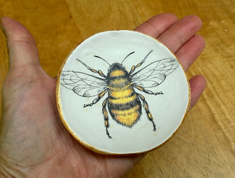 Op de afbeelding: Een wit keramiek sieradenschotel met een gouden rand heeft een handgeschilderde illustratie van een hommel met zwarte en gele strepen.