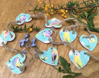 May include: A garland of blue heart-shaped ornaments with a string of twine. Each heart features a different colourful bird and floral design.