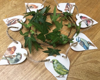 May include: A garland of white heart-shaped ceramic ornaments featuring different bird illustrations. The garland is strung with twine and includes a variety of birds, such as a blue tit, a greenfinch, and a robin.