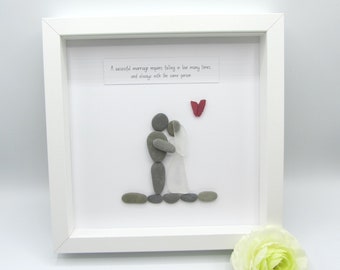 May include: A framed artwork featuring a pebble art depiction of a couple embracing. The figures are made of gray pebbles, with a white dress and veil for the bride. A red heart and a quote about marriage are included.
