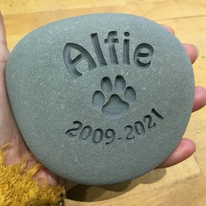 May include: A gray stone with the name "Alfie" engraved on it, along with a paw print and the dates "2009-2021".