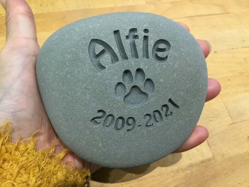 May include: A gray stone with the name "Alfie" engraved on it, along with a paw print and the dates "2009-2021".