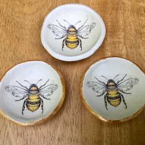 May include: Three white ceramic trinket dishes with gold rims, each featuring a black and yellow bee illustration.