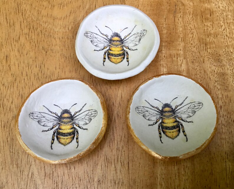 May include: Three white ceramic trinket dishes with gold rims, each featuring a black and yellow bee illustration.