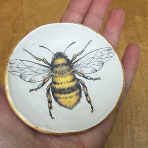 Op de afbeelding: Een wit keramiek sieradenbakje met een gouden rand heeft een handgeschilderde illustratie van een hommel met zwarte en gele strepen.